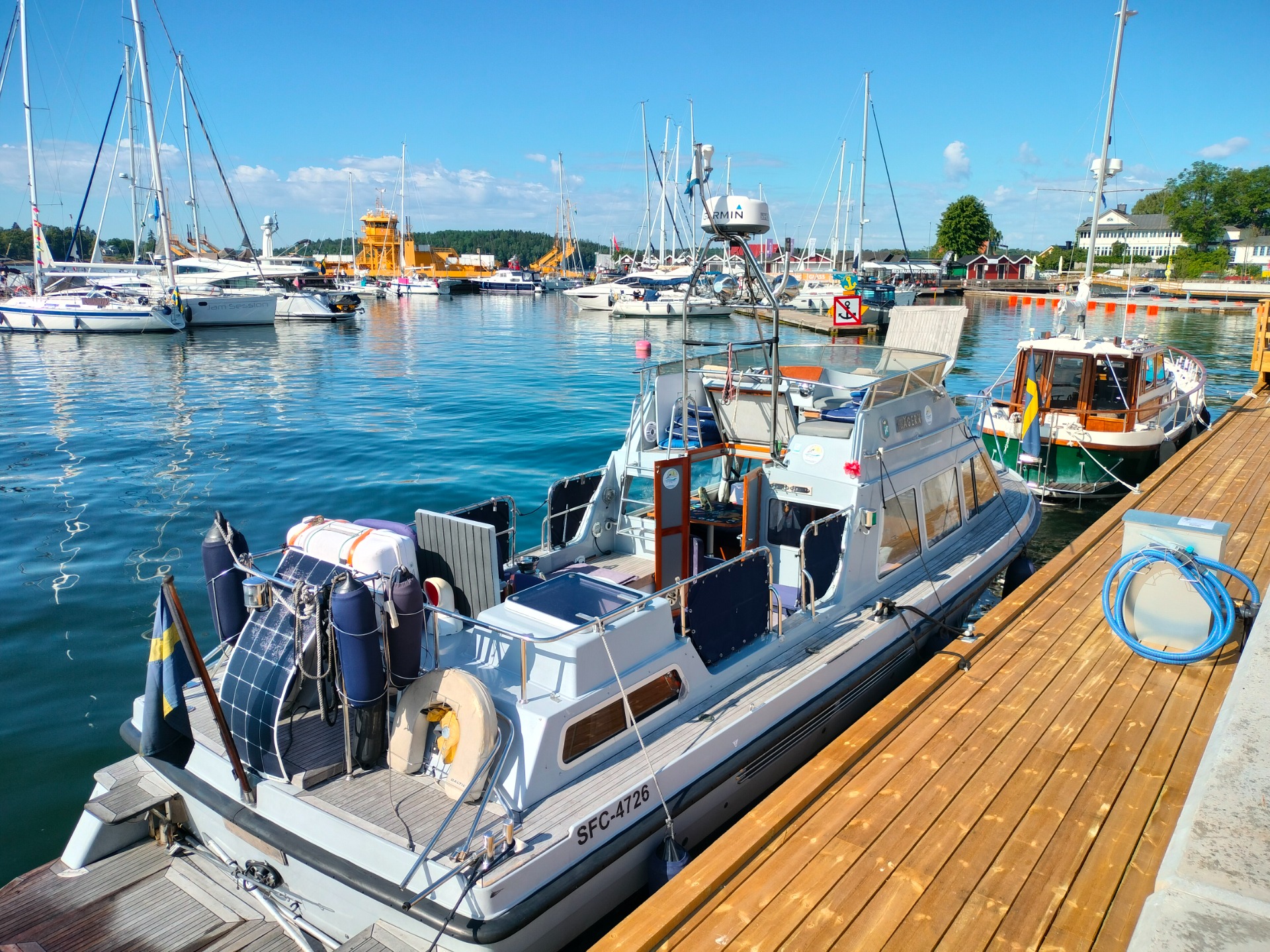 Grå  charterbåt vid brygga med segelbåtar i hamnen, blå himmel och spegelblankt vatten.
